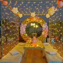 I created this magical bride-to-be party on a terrace, perfect for an evening celebration. The setup includes a canopy of fairy lights, low seating with cushions, and a circular balloon arch in pastel shades. A 'Bride to Be' neon sign serves as the centerpiece for photos.