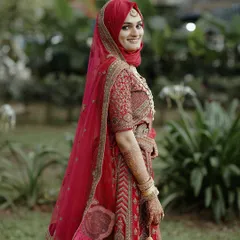 A classic bridal portrait. The bride looks over her shoulder with a beautiful smile, her stunning red lehenga standing out against the lush green background.