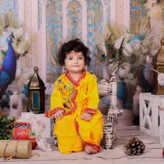 A baby dressed in a yellow kurta sits calmly on a small chair in our Janmashtami setup. The peacock feathers and painted pots add a traditional touch.