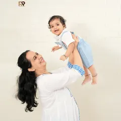 A heartwarming portrait of a mother lifting her smiling baby. The simple white brick background keeps the focus on their beautiful interaction and loving bond.