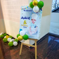 A close-up of a personalized welcome board for a first birthday party. The sign features the child's photo and is decorated with a small balloon bunch.