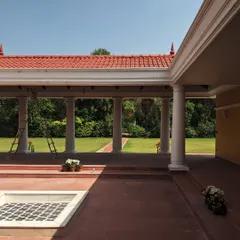 A view from the veranda of an upcoming farmhouse, where traditional red stone flooring and a tiled roof meet the green landscape. The design creates a classic, lived-in charm that respects Indian architectural heritage.