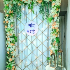 An elegant and simple backdrop for an at-home 'Godh Bharai' ceremony. The design features a lattice frame decorated with peach and white flowers, with fairy lights woven in for a gentle glow.