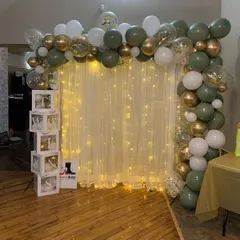 An elegant and simple backdrop solution for any event. This design features a curtain of warm fairy lights behind a sheer white drape, framed by an organic balloon garland in olive green, white, and gold.