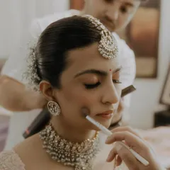 A behind the scenes moment, perfecting a bride's look. The hairstyle is a neat low bun with baby's breath, complementing her intricate jewelry and soft makeup.