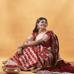A full-length portrait that celebrates the rich color and texture of a traditional red Banarasi saree. The relaxed, seated pose and genuine smile give the image a natural and joyful feel, highlighting the beauty of the garment in a relatable way.