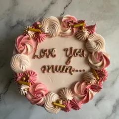 An overhead view of a beautiful Mother's Day cake. It features a message of "Love You Mum" written in edible gold, surrounded by a wreath of pink and white meringue kisses.