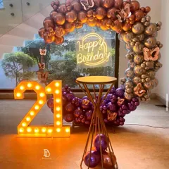 A chic 21st birthday party featuring a circular balloon arch with a mix of metallic rose gold, silver, and purple balloons. The look is completed with large '21' marquee numbers and a 'Happy Birthday' neon sign, creating a perfect photo spot.