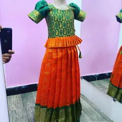 This is a classic kids' Paavada blouse set in a bright orange and green silk combination. The top has a peplum style and puff sleeves, all made with very good stitching for a clean look.