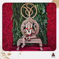 A modern and elegant Ganesha setup. The brass idol is placed against a backdrop of vibrant red flowers and green foliage, with a custom monogram above, blending tradition with contemporary style.