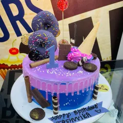 Celebrating a first birthday with a colorful and exciting drip cake. This custom piece combines purple and blue layers and is loaded with fun toppings like sprinkle donuts, an ice cream cone, Oreo cookies, and chocolate wafer rolls.