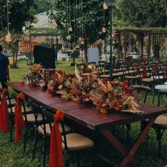 An overview of the guest seating arrangement for an intimate outdoor wedding. A long wooden table with a lush floral runner and hanging fairy lights creates a communal and magical dining experience under the open sky.