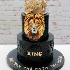 A cake fit for a king to celebrate an 80th birthday. This two-tiered design in black ganache features a hand-painted lion, intricate stencil work, and a bold golden crown topper.