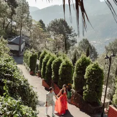 A high-angle shot of a couple walking down a beautiful, tree-lined path in the hills of Mussoorie. The vibrant colors of their outfits pop against the lush greenery.