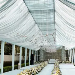 The aisle for the Anand Karaj ceremony, lined with white and blue flowers leading to the main stage. The ceiling is draped in soft, translucent fabric with hundreds of tiny hanging bells.
