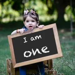 An adorable one year old girl peeking over a chalkboard sign that says "I am one". This is a classic and cute way to announce a first birthday.