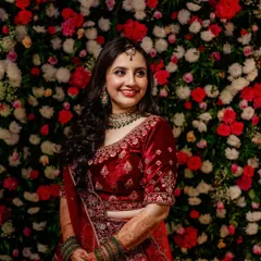 A happy bride against a floral backdrop. Her red velvet lehenga is paired with soft glam makeup and loose curls for a romantic reception look.