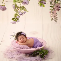 A baby in a purple wrap and headband sleeps in a bowl, with delicate lavender flowers hanging from above, creating a serene and color-themed scene.