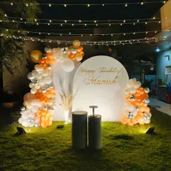 An elegant and simple garden birthday decoration with a white and orange balloon arrangement, a personalized backdrop, and string lights overhead.