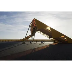 The dynamic, angular form of the footbridge creates a dramatic silhouette against the sky. The structure is designed to be experienced from multiple viewpoints, both by pedestrians and by drivers passing below.