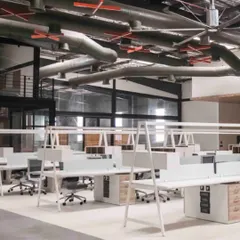 An aerial view of the open-plan workstation area at the DesignCo industrial office. The design utilizes the building's height and industrial character, with exposed MEP and large fans for air circulation.