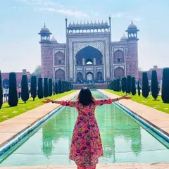 A joyful, open-armed pose in front of the main gate of the Taj Mahal, wearing a vibrant pink floral ethnic dress that complements the scene beautifully.