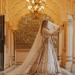 A bride in a magnificent shimmering lehenga stands in a grand palace corridor, the long veil adding to the drama and elegance.