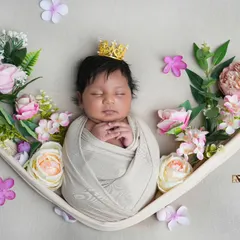 A newborn princess rests in a soft, cloth-lined hammock, crowned and surrounded by delicate pink and white flowers. This artistic setup creates a beautiful, fairytale-like portrait.