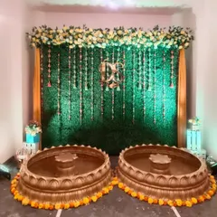 A Haldi ceremony setup with a green grass wall and hanging floral strings. Two large urlis are placed for the rituals, decorated with marigold garlands.