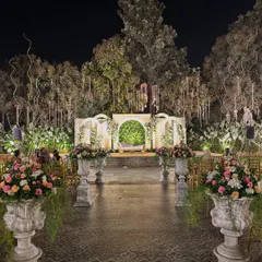 A wide view of an elegant outdoor night ceremony, with a minimalist white stage, large floral urns, and trees adorned with hanging lights.