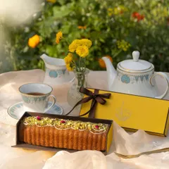 Our Pistachio Tea Cake, unboxed and ready to be enjoyed. This moist and flavourful cake is topped with pistachios and rose petals, presented alongside our elegant yellow gift box in a lovely garden tea party setting.