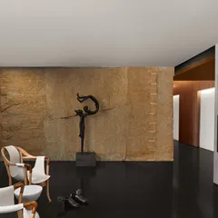 A detail shot of the passage at the Baid Residence, showing the juxtaposition of a rough, natural stone wall, the sleek, honed black granite floor, and the smooth finish of designer chairs.