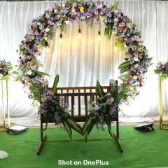 This naming ceremony stage features a beautiful cradle decorated with a mix of colorful flowers. The circular floral arch behind it is filled with roses, carnations, and chrysanthemums, set against a simple white curtain.