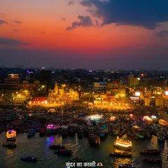 An aerial view of the Kashi ghats during a vibrant sunset. The sky is painted in hues of orange and purple, reflecting over the Ganga, where countless boats and people have gathered for the evening Aarti.