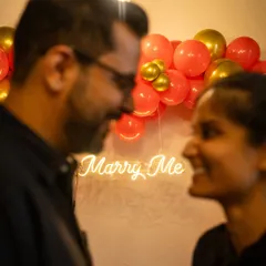 A close-up shot capturing the intimacy of a proposal. The soft glow of the 'Marry Me' neon sign creates a romantic and personal moment between the couple.