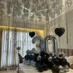A wide shot of the 30th birthday surprise room decor. This shows the full effect of the silver balloons on the ceiling with black ribbons, and the large balloon arrangement on the bed.
