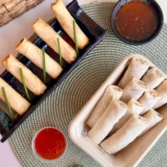 These are my signature spring rolls, available as a ready-to-cook option. The image displays them both frozen and perfectly fried, served with sweet chili sauce, showcasing the convenience of my cook-and-serve range.