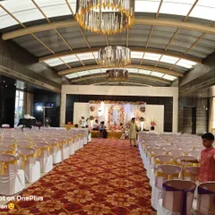 This wide shot shows a complete hall setup for a wedding ceremony. You can see the main stage decor, the aisle carpet, and the neat arrangement of chairs with covers, all ready for the guests to arrive.