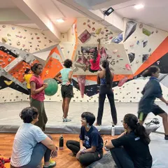The gym buzzes with energy during a women's climbing workshop. While some climbers are on the wall, others rest, watch, and cheer each other on, creating a dynamic and supportive atmosphere.