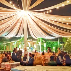 A wide-angle view of an outdoor reception at night, with guests seated under a large tent canopy illuminated by hanging bulb lights.