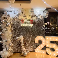 Celebrating a silver jubilee in style. This 25th-anniversary decor features a shimmering silver sequin wall, a custom "Happy Anniversary" neon sign, and large light-up marquee numbers, all framed by an elegant white and silver balloon arrangement.