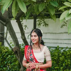 A beautiful portrait of the bride, Aishwarya, in her traditional red and white saree. Her happy, natural smile shines against the lush green backdrop.