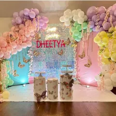 A vibrant butterfly-themed party featuring a stunning iridescent shimmer wall that catches the light beautifully. The setup is framed by a pastel rainbow balloon garland and includes themed plinths for the cake.