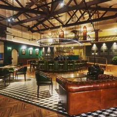 This wide shot of the KGA bar shows how different layers of light work together. Uplighting on the wooden ceiling trusses adds height and warmth, track lights highlight artwork on the green walls, and low-level table lighting creates intimate pockets within the larger space.