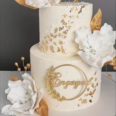 A vision in white and gold for a beautiful engagement celebration. This two-tiered cake features large, handcrafted white sugar flowers, delicate gold leaf accents, and a custom gold topper, creating a look of pure luxury and romance.