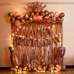 A very popular birthday decoration in a rose gold and gold theme. This setup includes a shiny foil curtain, 'Happy Birthday' letters, a balloon arch topped with a crown, and other fun balloons like a champagne bottle and a 'Love' sign.