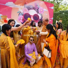 The groom is showered with flower petals by family and friends during the Haldi ceremony, with a colorful and artistic backdrop behind him.