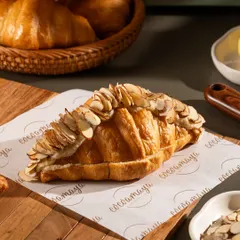 A flaky almond croissant, generously topped with sliced almonds. The lighting catches the golden-brown crust and buttery layers.