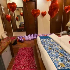 This romantic room setup features a thick pathway of red rose petals leading to the bed. The room is filled with red heart-shaped foil balloons and bunches of regular balloons hanging from the ceiling to create a classic and passionate atmosphere.