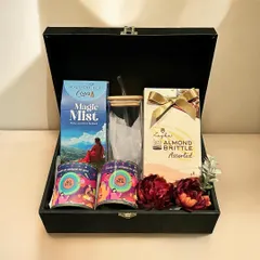 A premium Diwali hamper presented in a black wooden box, containing artisanal snacks, almond brittle, a glass tumbler, and aromatic mist for a complete sensory experience.